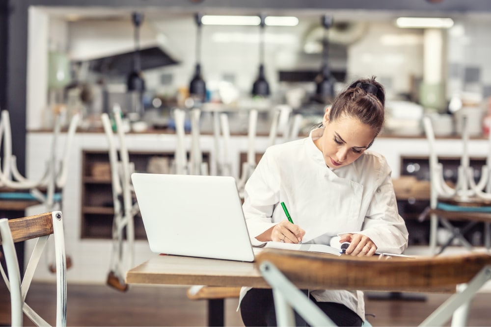 Female restaurant chef sits at the table after hours doing her bookkeeping in front of an open laptop